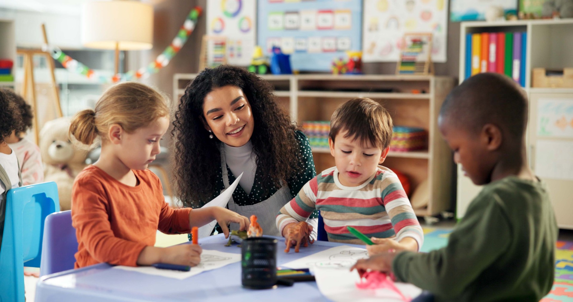A teacher in a classroom working with three students, each of which are coloring and have toy animals.